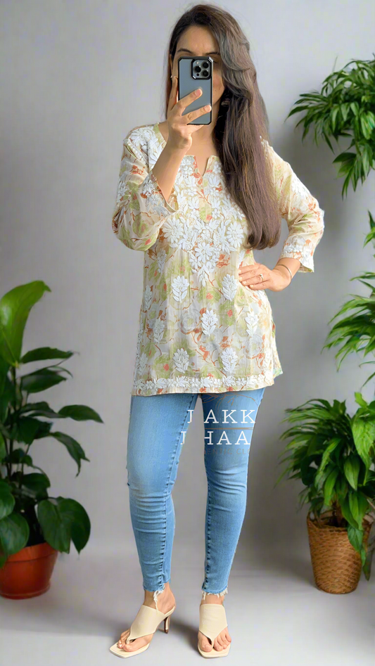 Person taking a mirror selfie wearing a floral chikankari top and blue jeans in a room with plants.