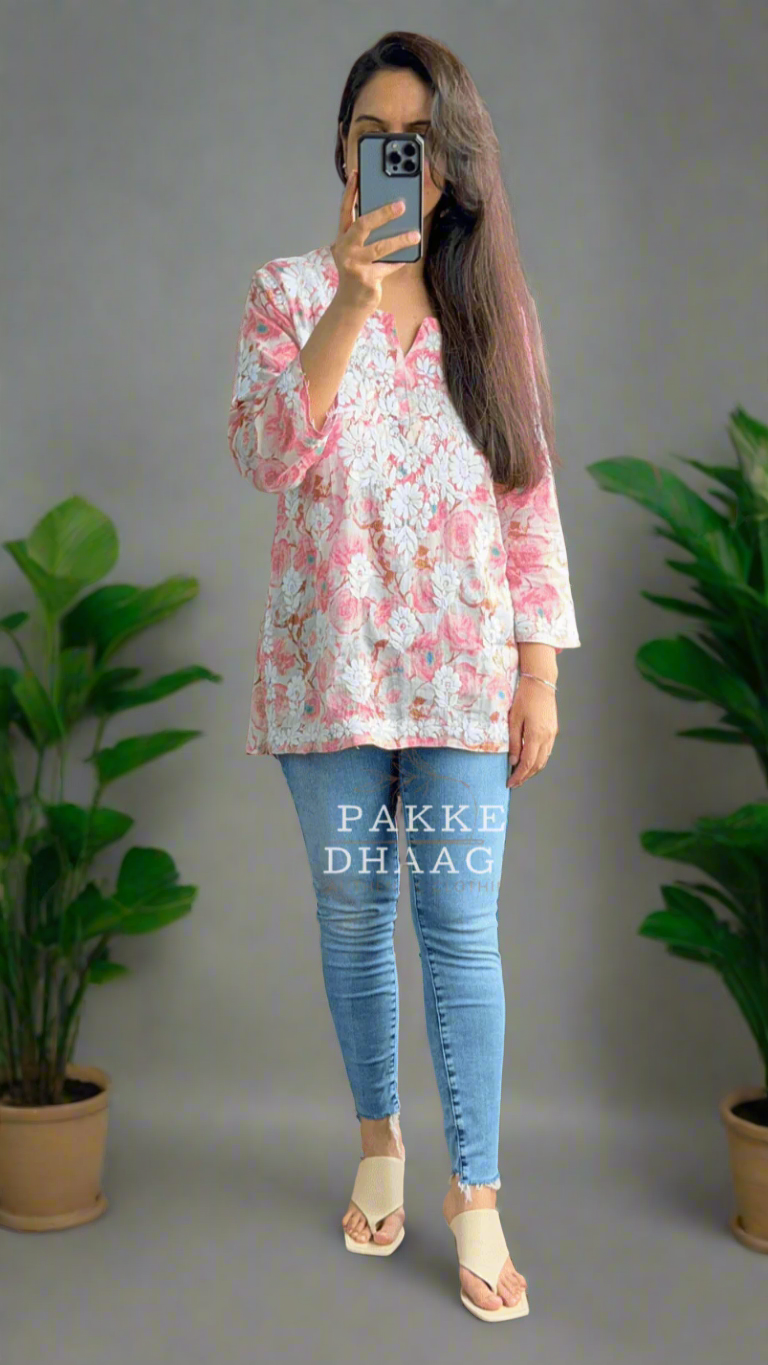 Person taking a mirror selfie wearing a floral pink chikankari top and blue jeans in a room with plants.