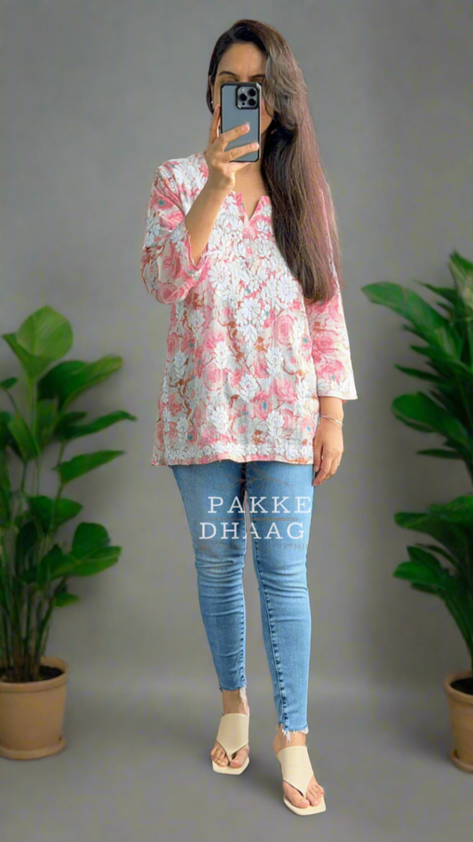 Person taking a mirror selfie wearing a floral pink chikankari top and blue jeans in a room with plants.