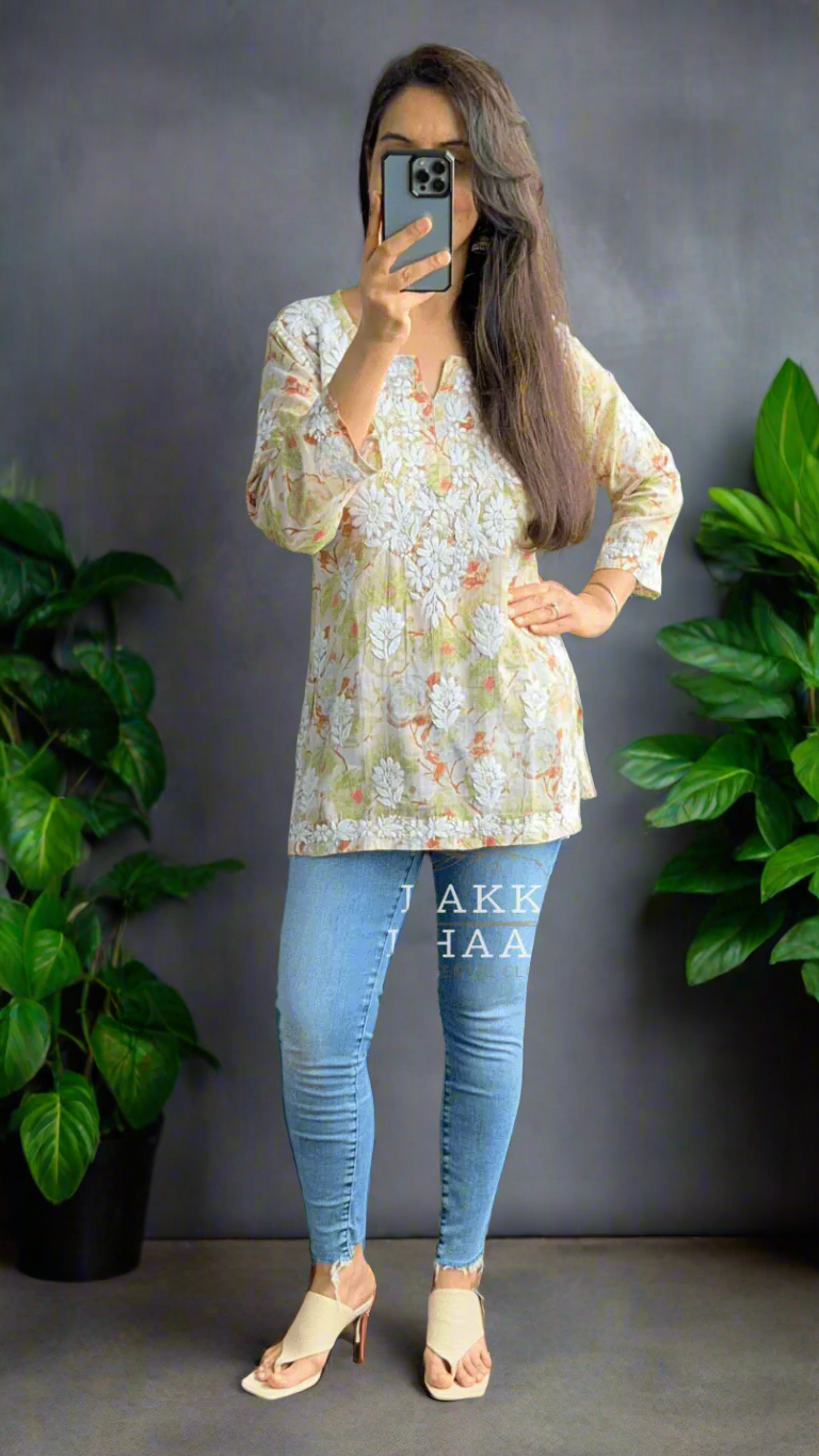 Woman taking a mirror selfie wearing a floral chikankari top and blue jeans in an indoor setting with plants.