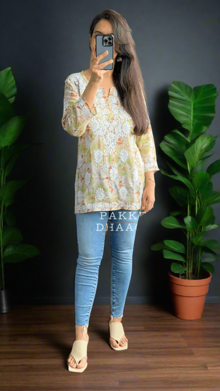 Woman taking a mirror selfie wearing a floral printed chikankari top and blue jeans in a room with plants.