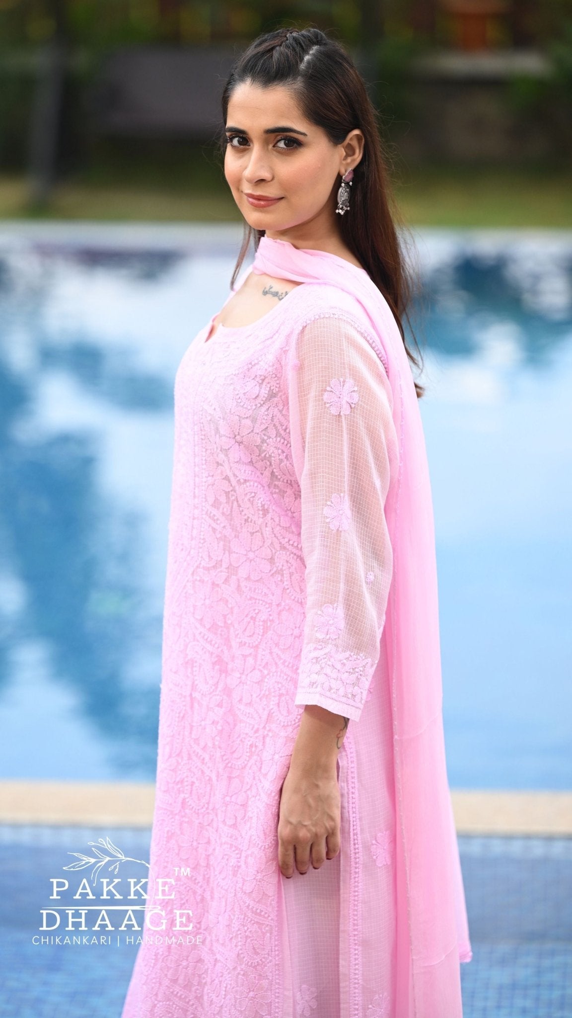 A woman wearing a light pink chikankari embroidered kurta with a matching sheer dupatta, standing outdoors near a swimming pool. The brand name 'PAKKE DHAAGE' is visible as a watermark in the bottom-left corner.