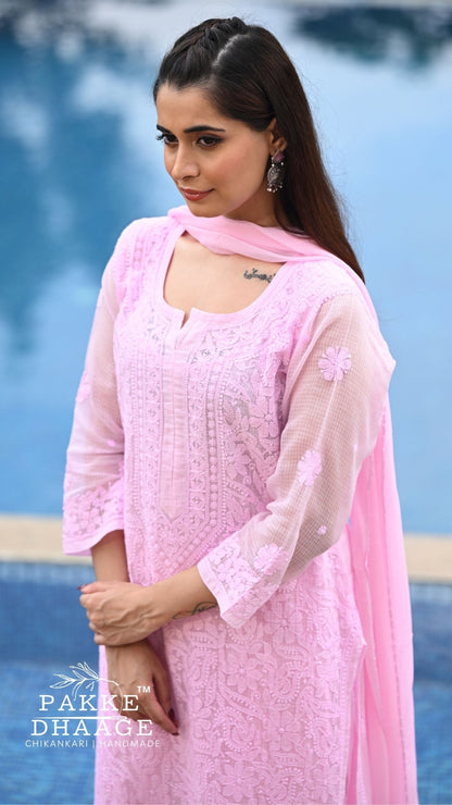 A woman wearing a light pink chikankari kurta set with a matching sheer dupatta, standing outdoors near a blue pool. The brand name 'Pakke Dhaage' is visible as a watermark in the bottom-left corner.