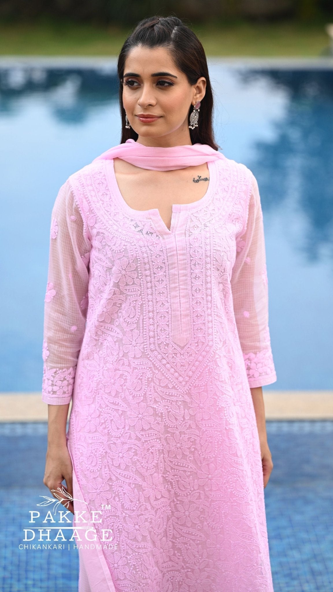 A woman wearing a light pink chikankari embroidered kurta with a matching dupatta, standing in front of a swimming pool. The brand logo 'Pakke Dhaage' is visible in the bottom-left corner.