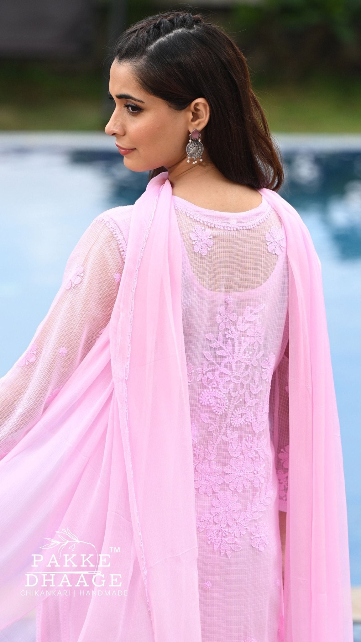 A woman wearing a light pink traditional outfit with floral chikankari and a matching sheer dupatta, photographed outdoors near a pool. The brand name 'PAKKE DHAAGE' is visible as a watermark in the lower-left corner.