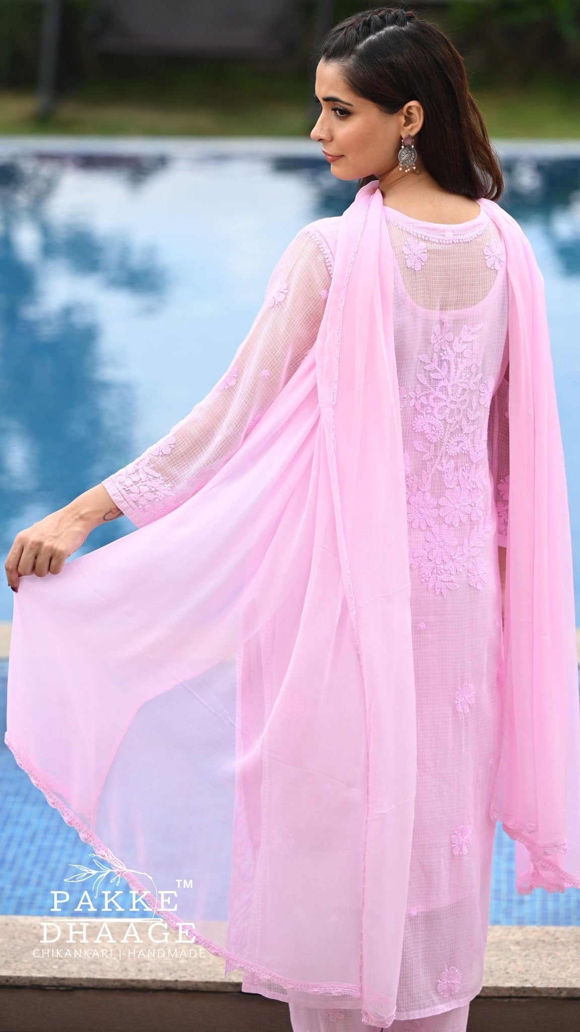 A woman wearing a light pink chikankari outfit with a matching sheer dupatta, standing beside a swimming pool. The outfit features floral embroidery on the back and sleeves.