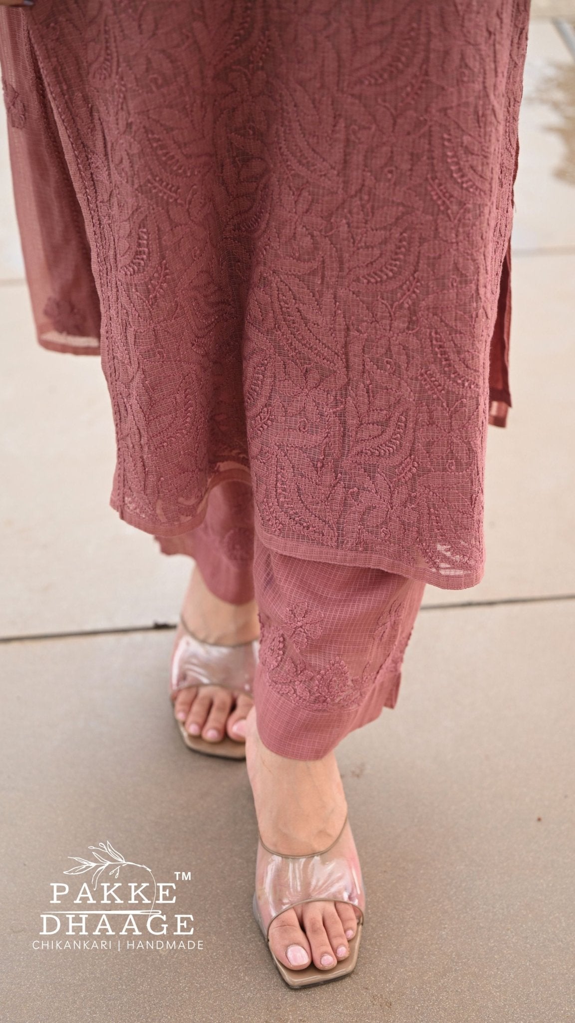 A close-up of a person wearing a dusty rose pink chikankari embroidered kurta and matching trousers with metallic pink sandals, standing on a concrete surface. The brand logo 'PAKE DHAAGE' is visible in the bottom-left corner.