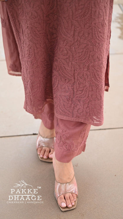 A close-up of a person wearing a dusty rose pink chikankari embroidered kurta and matching trousers with metallic pink sandals, standing on a concrete surface. The brand logo 'PAKE DHAAGE' is visible in the bottom-left corner.