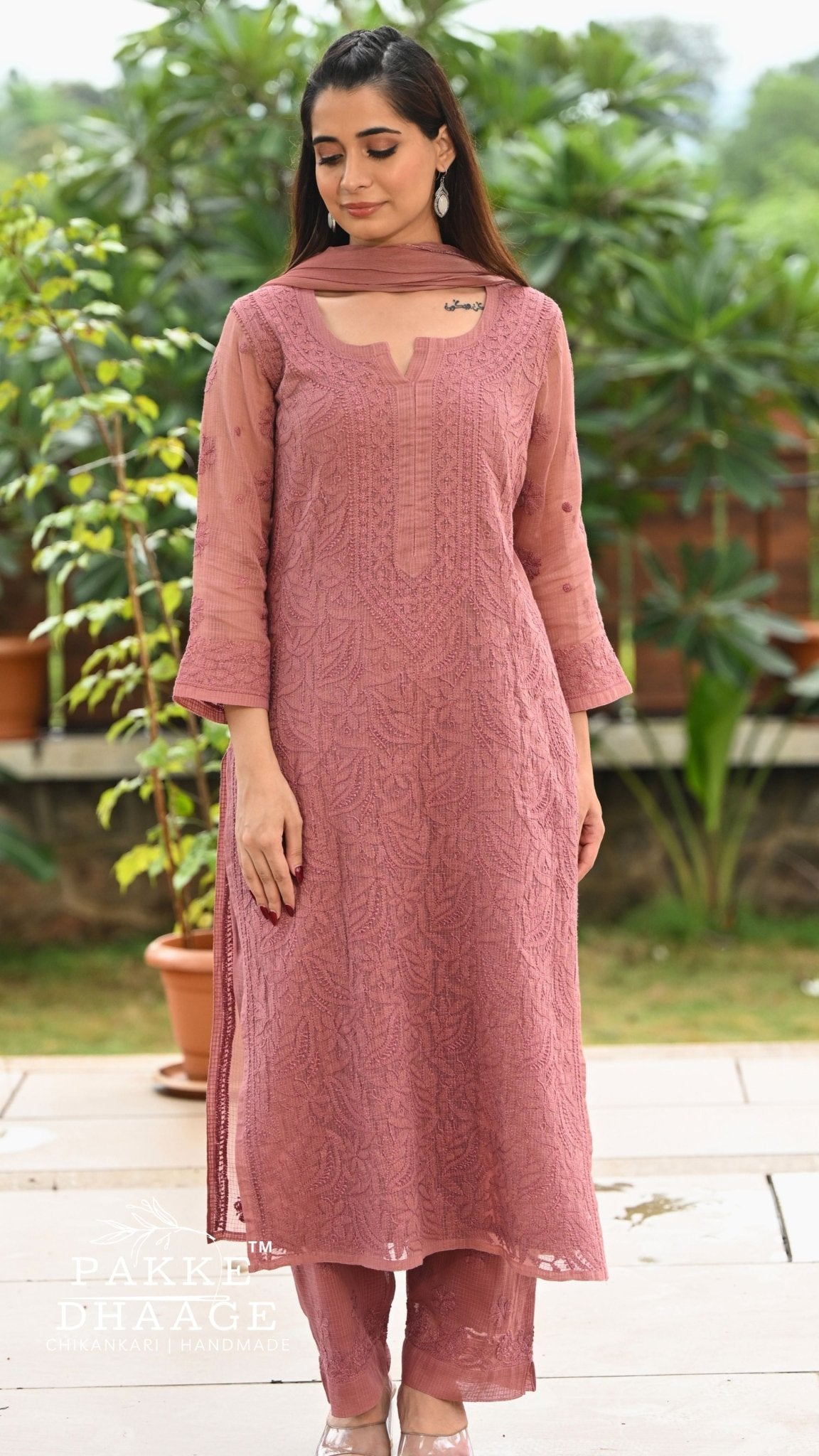 A woman modeling a chikankari embroidered kurta set with matching pants and dupatta in an outdoor garden setting. The kurta features intricate floral embroidery and a keyhole cutout at the neckline.