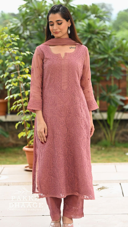 A woman modeling a chikankari embroidered kurta set with matching pants and dupatta in an outdoor garden setting. The kurta features intricate floral embroidery and a keyhole cutout at the neckline.