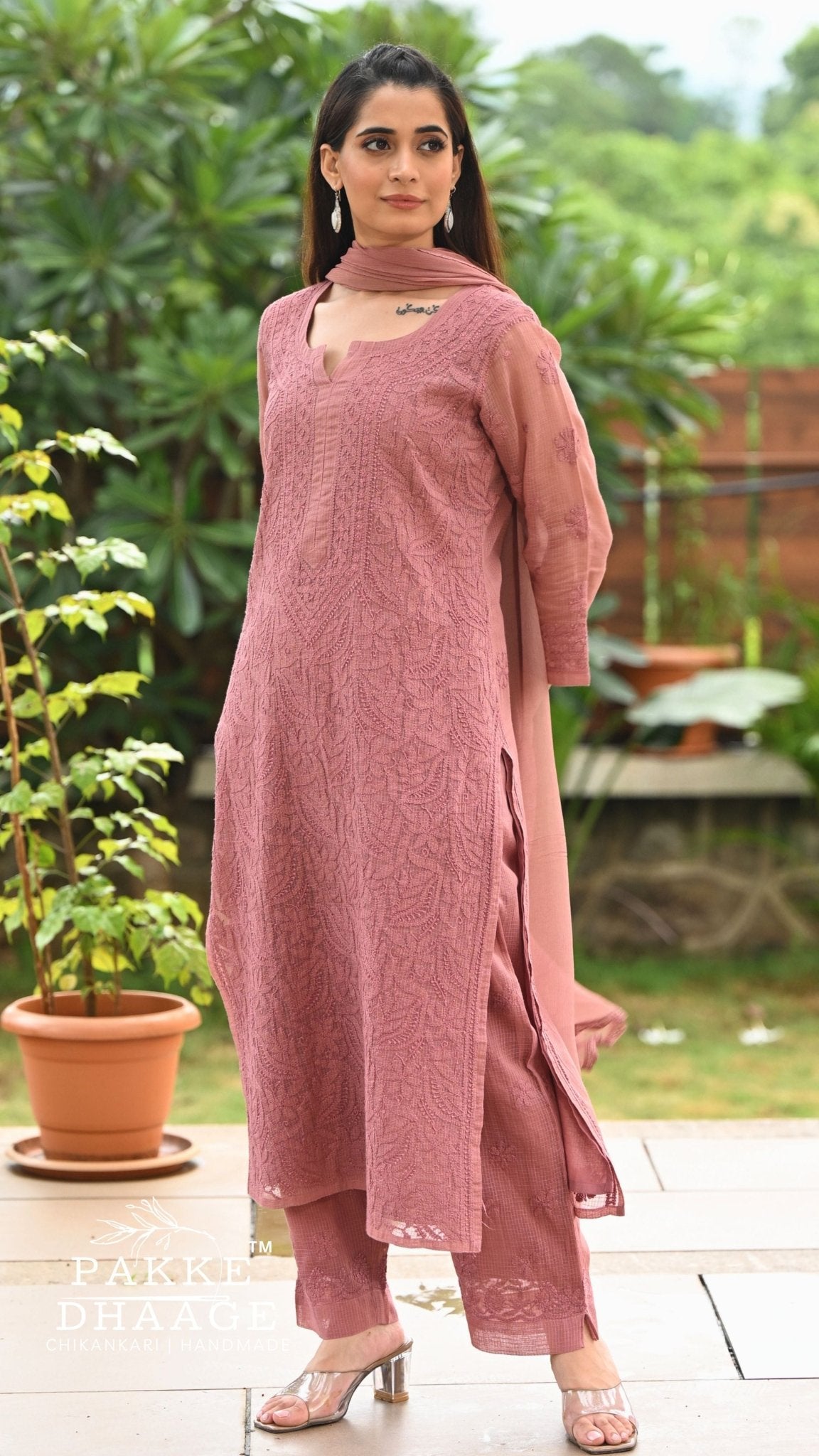 A woman modeling a chocolate chikankari ethnic three-piece suit with a matching dupatta in an outdoor garden setting. The set includes a kurta, trousers, and a long dupatta.