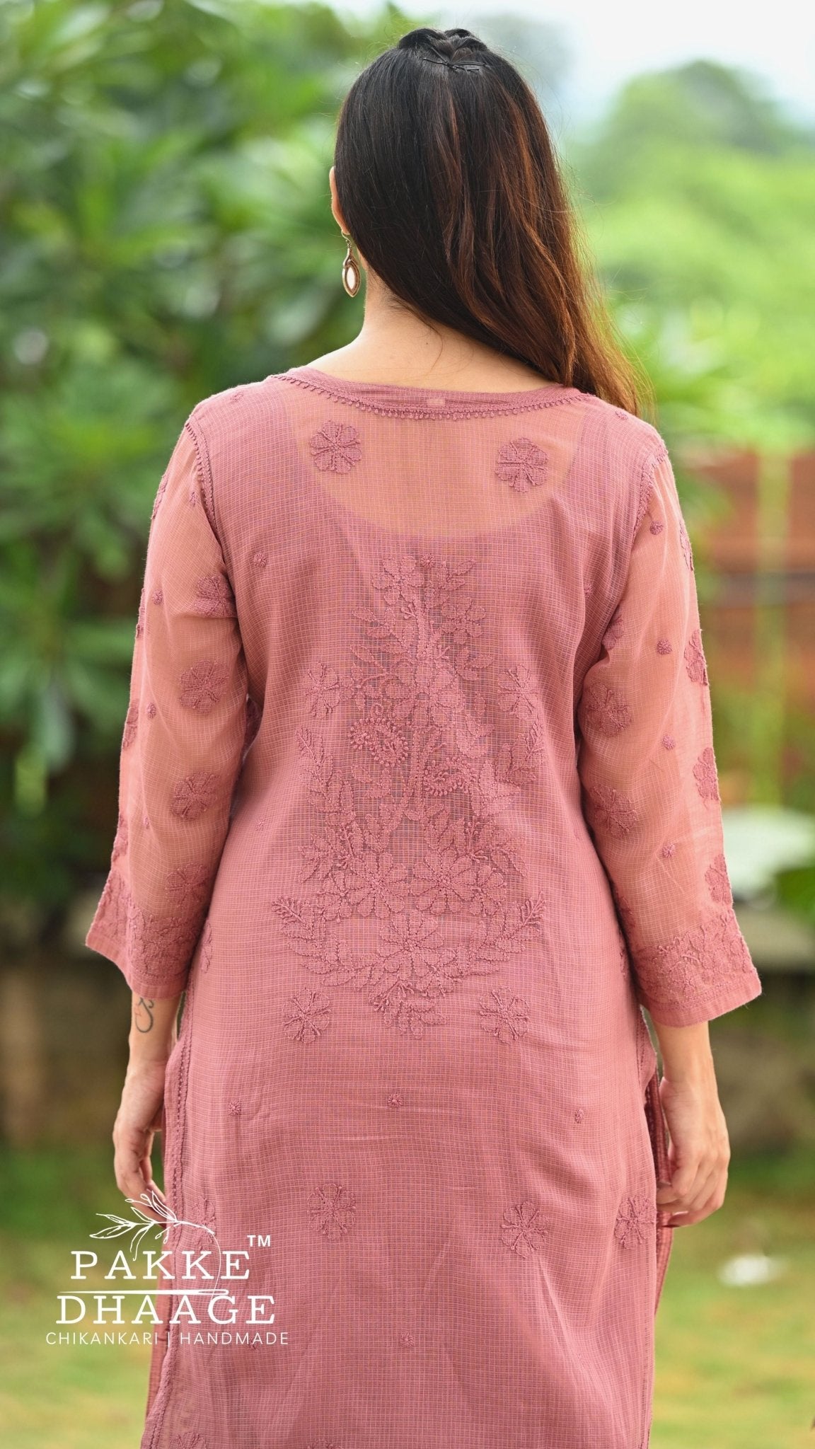 A woman viewed from behind wearing a dusty chocolate embroidered kurta with sheer sleeves, set against a blurred green garden background. The brand name 'Pakke Dhaage' is visible as a watermark in the bottom left corner.