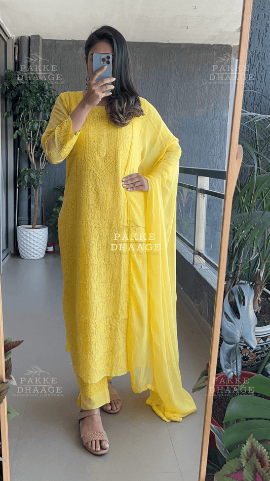 Person wearing a yellow chikankari outfit, standing in a room with plants and a mirror.
