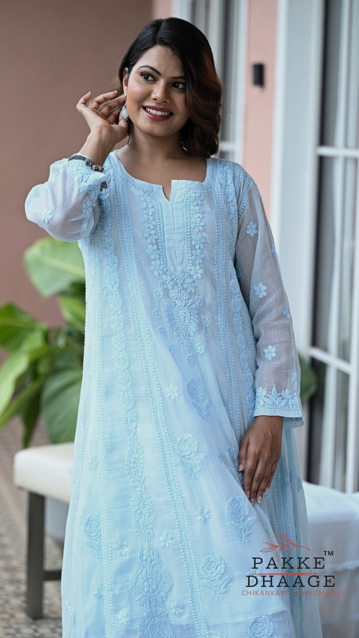 A woman wearing a light blue embroidered chikankari kurta with a matching dupatta, standing in front of a window. The brand name 'PAKKE DHAAGE' is visible as a watermark in the bottom right corner.