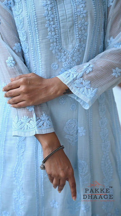 A close-up of a person wearing a light blue chikankari outfit with sheer sleeves and floral embroidery, showing both hands and part of the lower body. The brand name 'PAKKE DHAAGE' is visible in the bottom right corner.