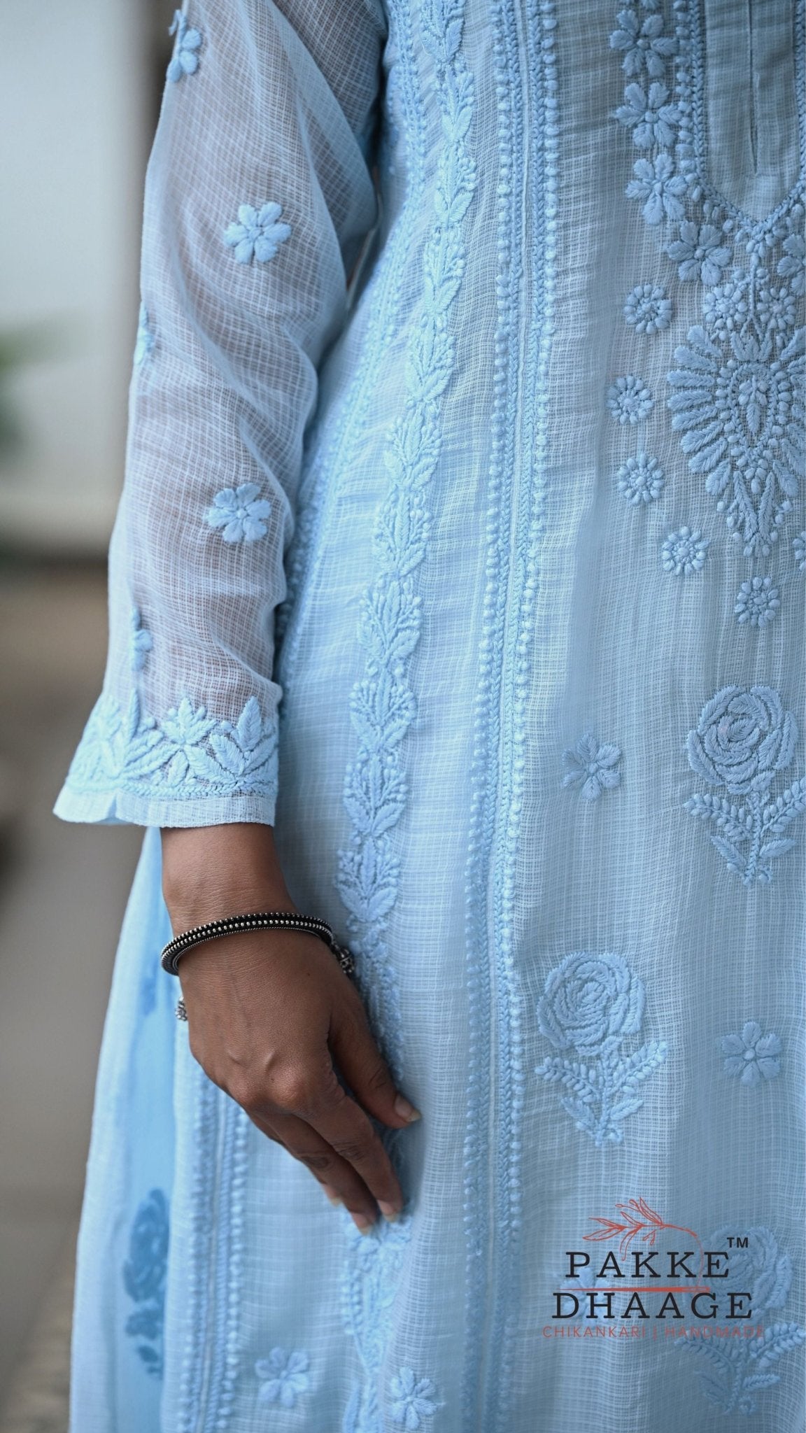 A close-up of a light blue embroidered chikankari dress with sheer sleeves and floral embroidery, worn by a model whose hand and arm are visible. The brand name 'PAkke DHAAGE' is printed on the bottom right.