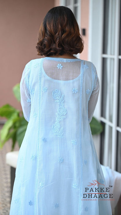 A woman viewed from behind wearing a light blue chikankari kurta with sheer mesh back panel and floral embroidery, standing indoors near a window. The brand name 'PAKKE DHAAGE' is visible as a watermark/logo at the bottom right.