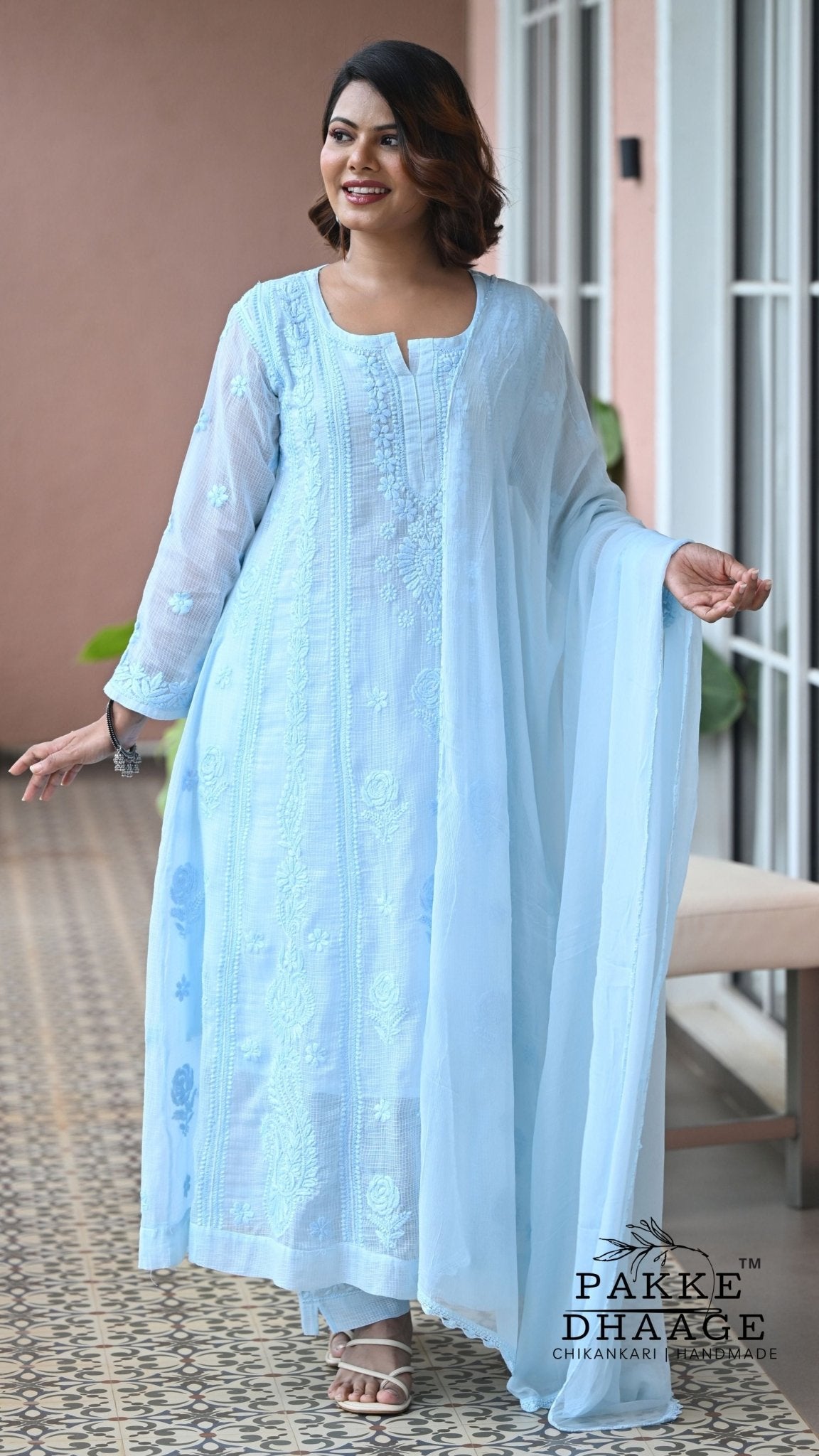 A woman modeling a light blue Chikankari embroidered kurta set with a matching dupatta, standing on a patterned tile floor in front of a building entrance. The brand name 'PAKKE DHAAGE' is visible as a watermark in the bottom right corner.