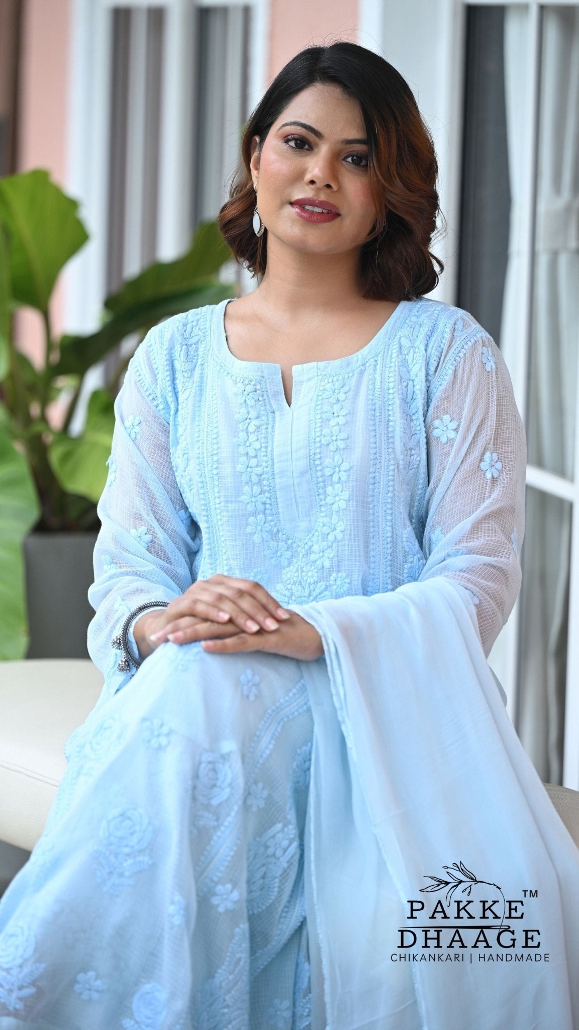 A woman wearing a light blue embroidered chikankari outfit with sheer sleeves and a matching dupatta, seated indoors. The brand name 'PAKKE DHAAGE' is visible as a watermark/logo in the bottom right corner.
