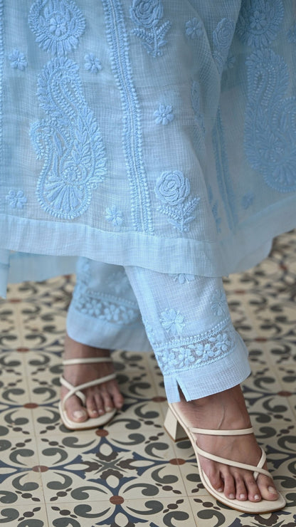 A close-up of a person wearing a light blue chikankari embroidered traditional outfit with matching pants and beige strappy sandals, standing on a patterned tile floor.