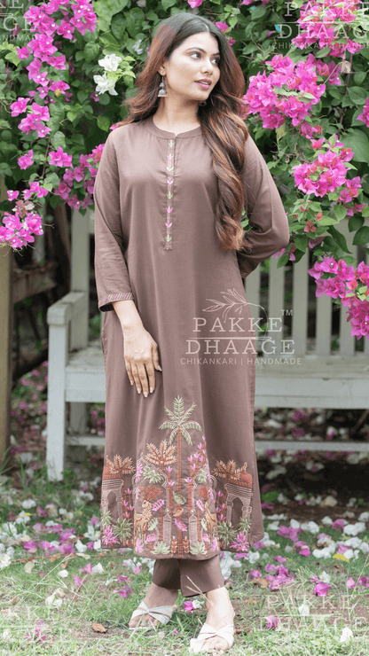 Woman in a brown embroidered suit standing in a garden with pink flowers and a bench.