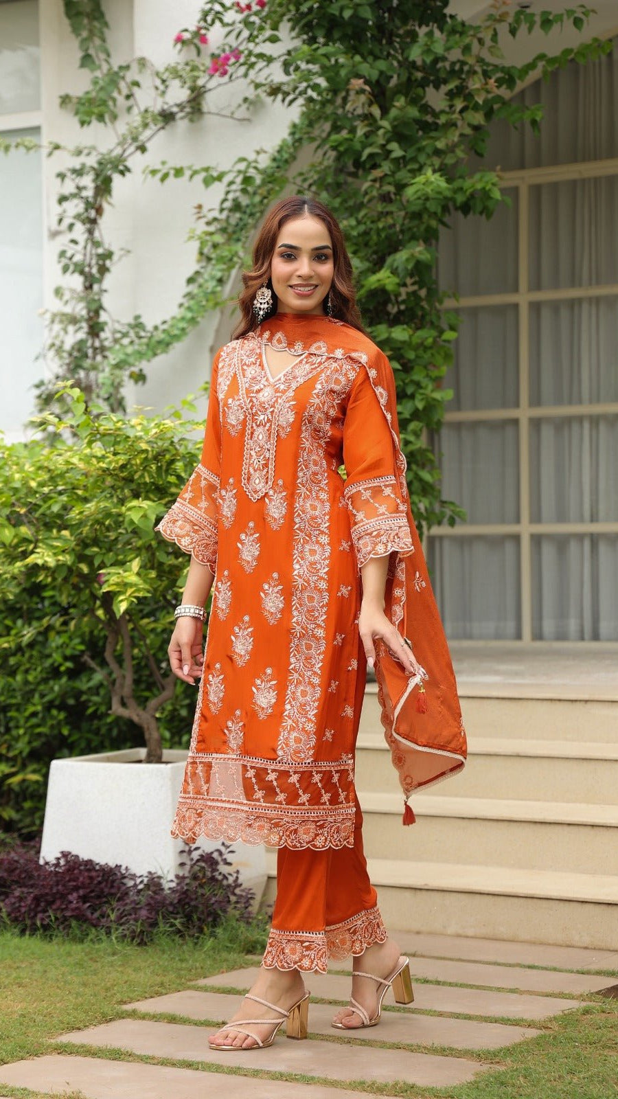 Woman in an orange traditional outfit standing outdoors with greenery and a building in the background