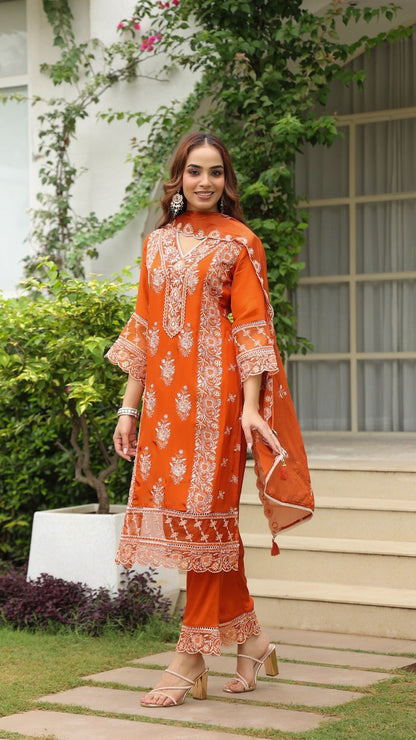 Woman in an orange traditional outfit standing outdoors with greenery and a building in the background