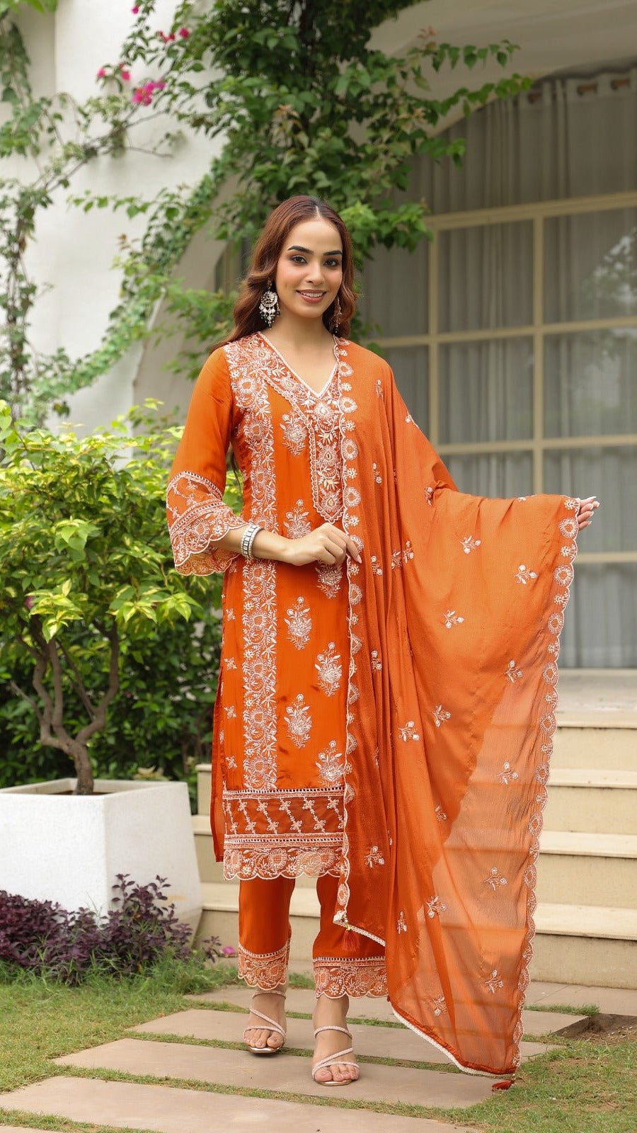 Woman in an orange traditional outfit standing outdoors with greenery and a building in the background.