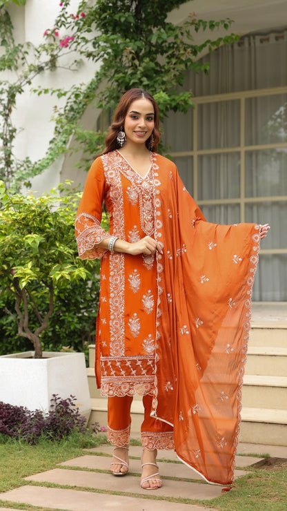 Woman in an orange traditional outfit standing outdoors with greenery and a building in the background.
