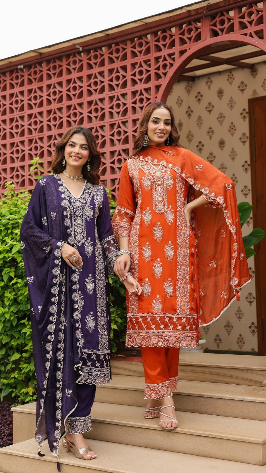 Two women in traditional outfits standing on steps with a decorative wall in the background.