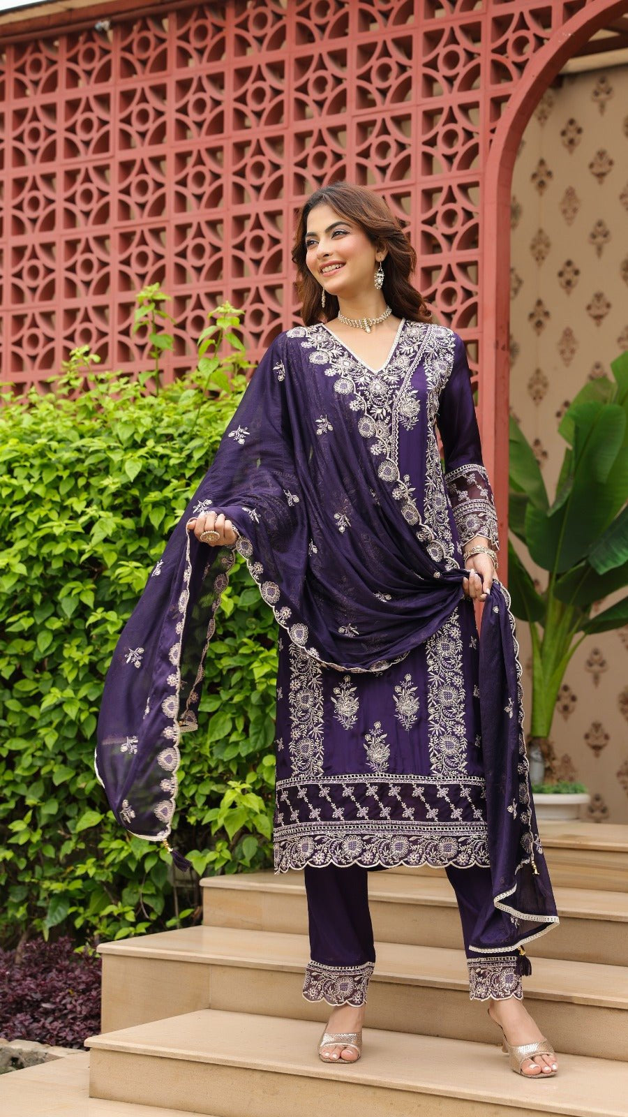 Woman in a purple traditional outfit standing on steps with decorative wall and plants in the background