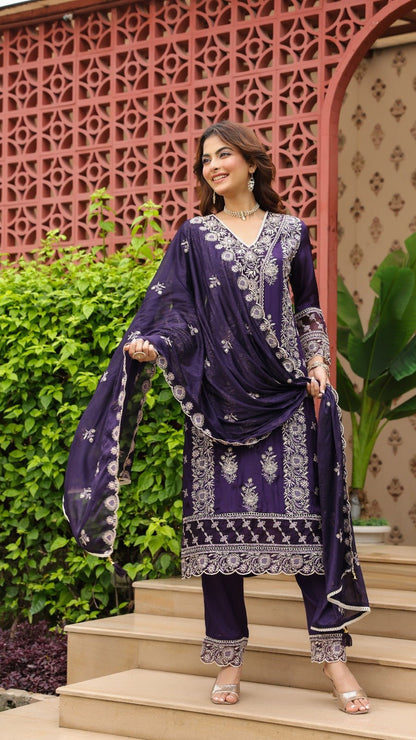 Woman in a purple traditional outfit standing on steps with decorative wall and plants in the background