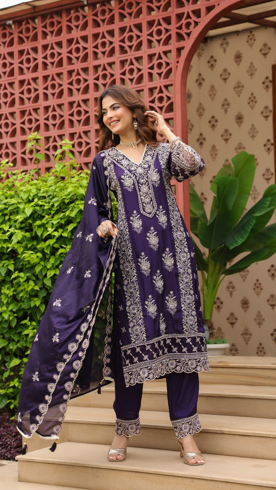Woman in a purple traditional outfit standing on steps with decorative wall and plants in the background