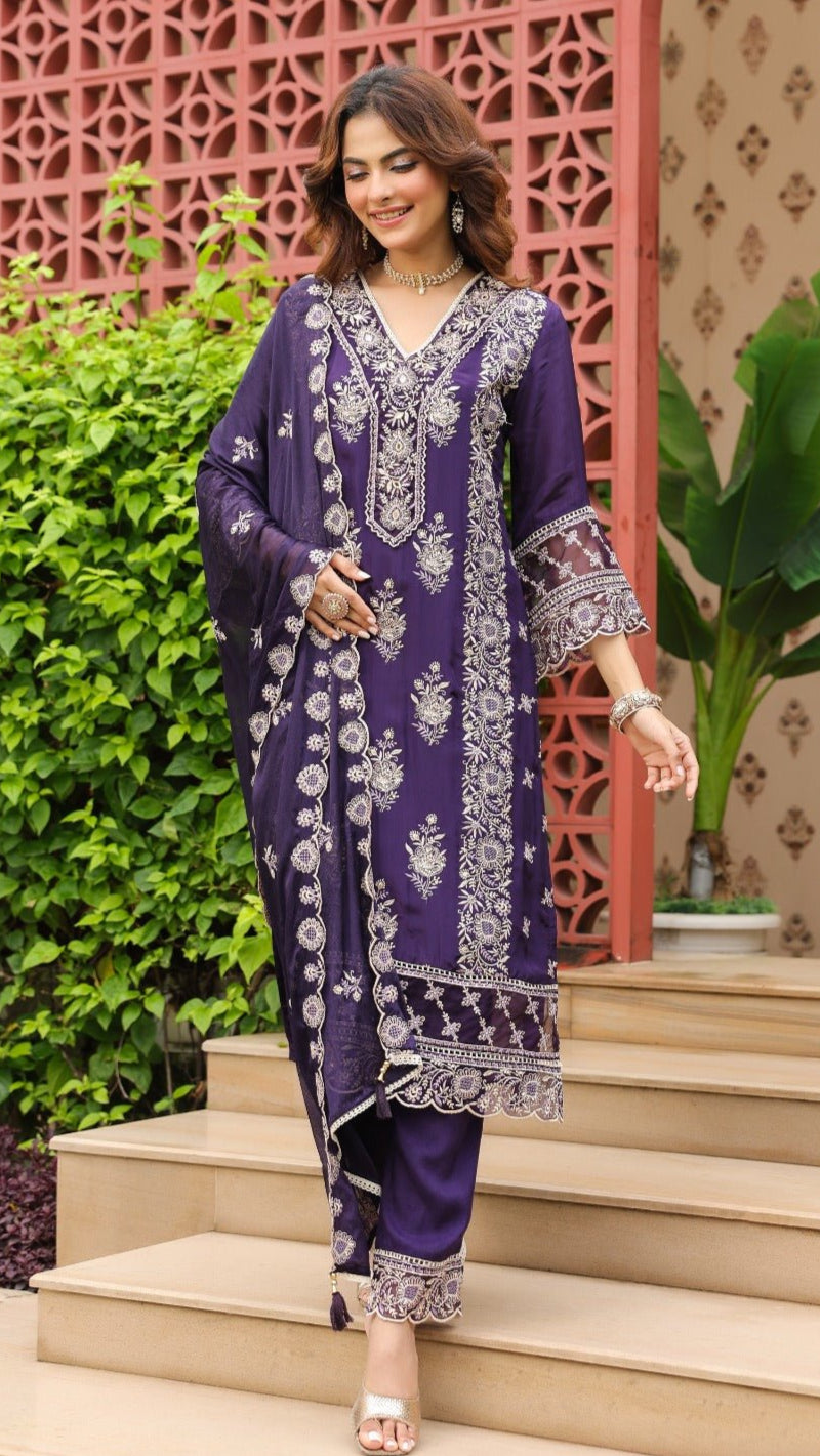 Woman in a purple traditional outfit standing on steps with decorative wall and plants in the background