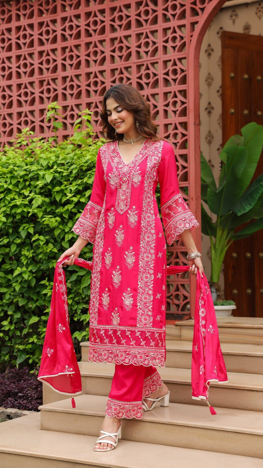 Woman in a bright pink traditional outfit standing on steps with a decorative pink archway in the background.