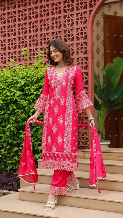 Woman in a bright pink traditional outfit standing on steps with a decorative pink archway in the background.