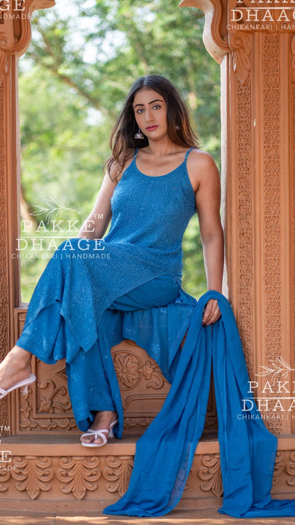 Woman in a blue traditional outfit standing in front of ornate wooden doors with 'Pakke Dhaag' branding.