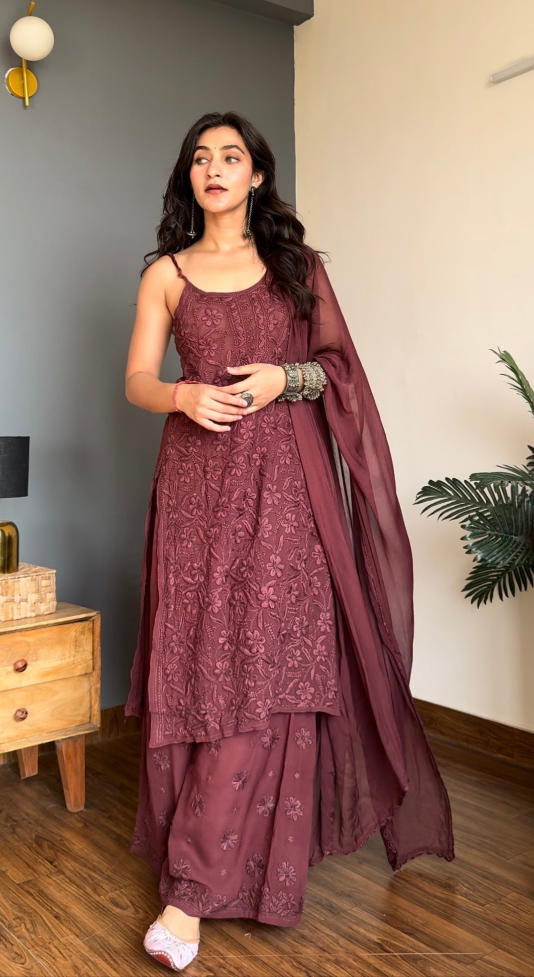 Woman in a maroon traditional chikankari kurta set outfit standing in a room with a lamp and plant.