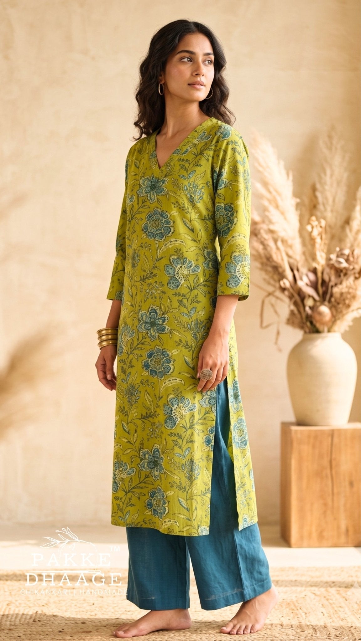 A woman modeling a lime green cotton kurta with blue floral block prints, paired with teal wide-leg pants, in a warm, rustic indoor setting. | Pakke Dhaage