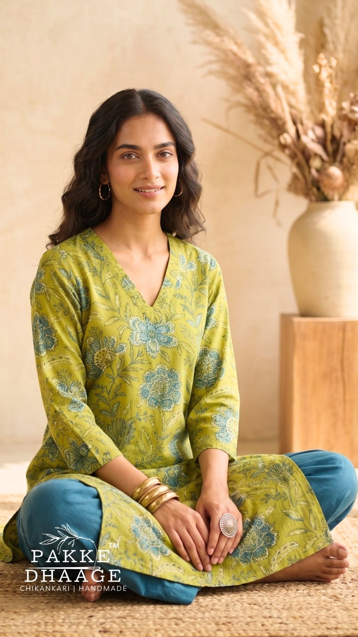 A woman sits cross-legged on a woven rug wearing a lime green printed kurta with blue pants. | Pakke Dhaage