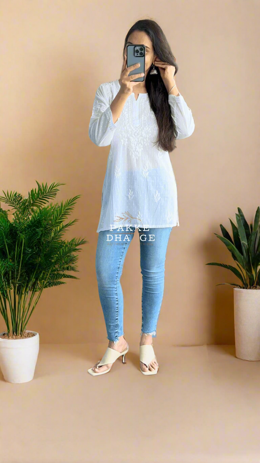 Person taking wearing a white chikankari backdrop top and jeans in an indoor setting with plants.