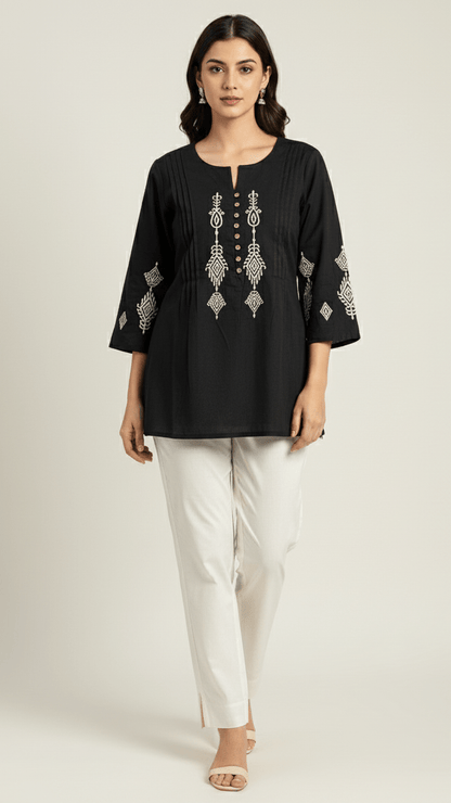 Woman wearing a black embroidered top and white pants on a light gray background