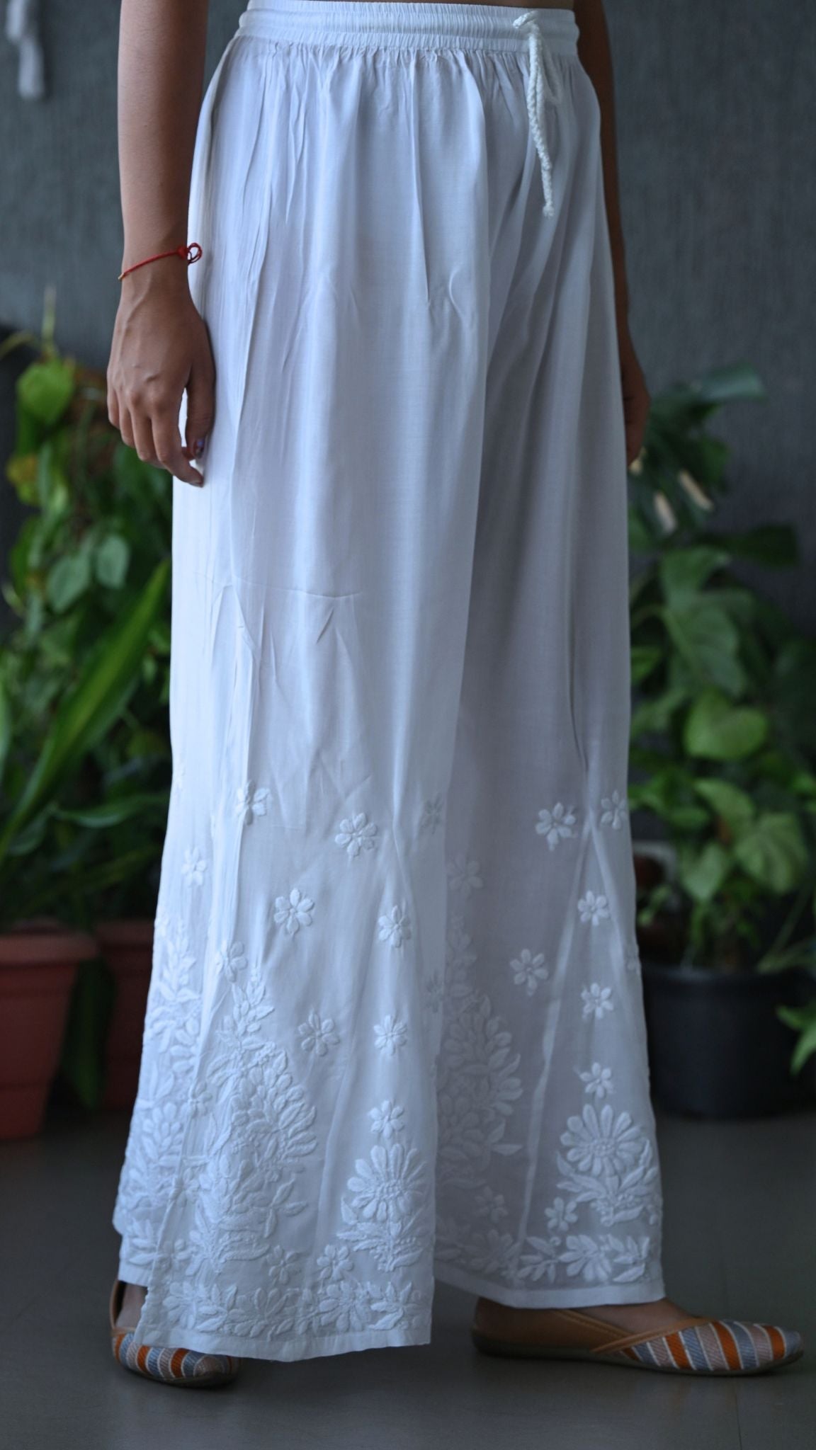 White wide-leg cotton maxi pants with floral chikankari embroidery near the hem, shown on a model in a styled indoor setting with potted plants in the background. | Pakke Dhaage