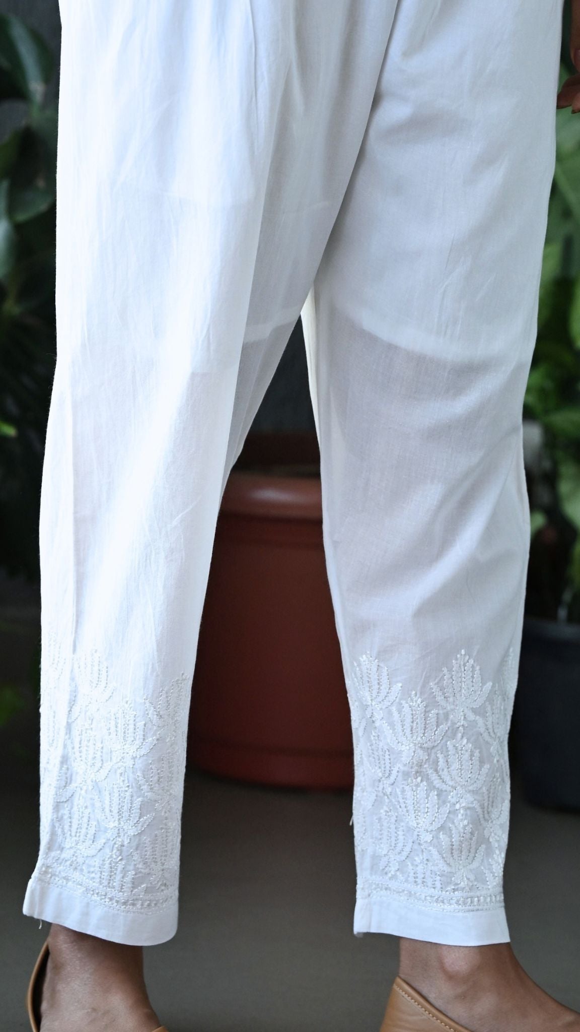 White cropped trousers with delicate floral embroidery at the hem, shown on a model standing outdoors. Handmade Chikankari | Pakke Dhaage