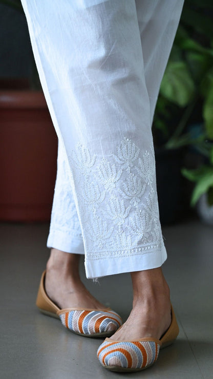 White embroidered pants with floral lace detailing at the hem, worn with tan flat shoes featuring orange, white, and gray striped uppers, shown on a person standing indoors. Handmade Chikankari | Pakke Dhaage