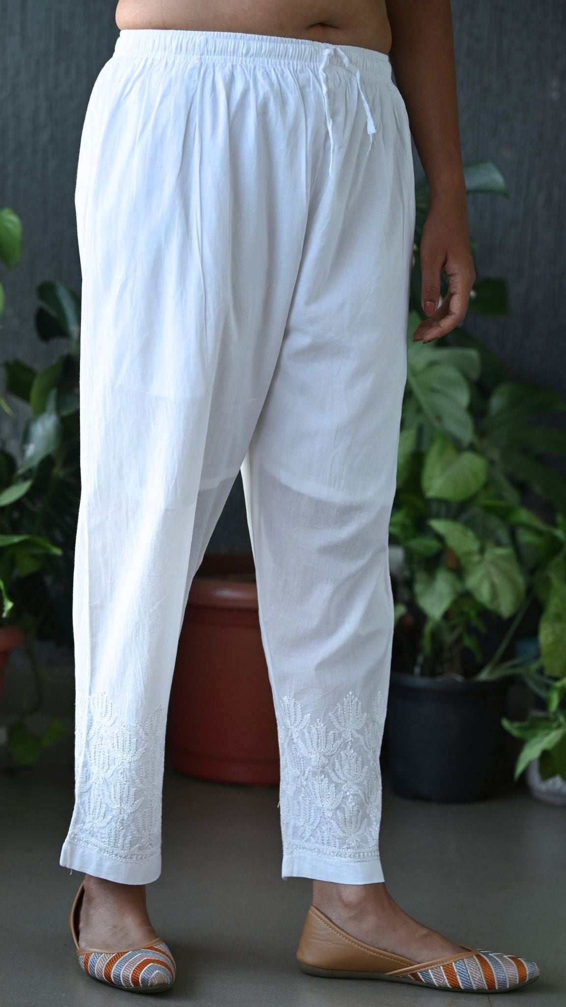 White cotton drawstring pants with delicate floral embroidery near the hem, shown on a model wearing flat shoes in front of a dark gray wall and potted green plants. | Handmade Chikankari | Pakke Dhaage