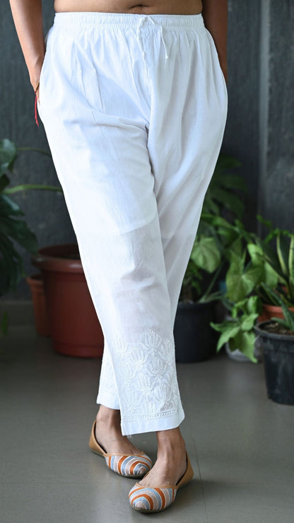 White cotton cropped pants with a drawstring waist and delicate floral embroidery near the hem, shown on a model wearing flat sandals in an indoor setting with potted plants. Handmade Chikankari | Pakke Dhaage