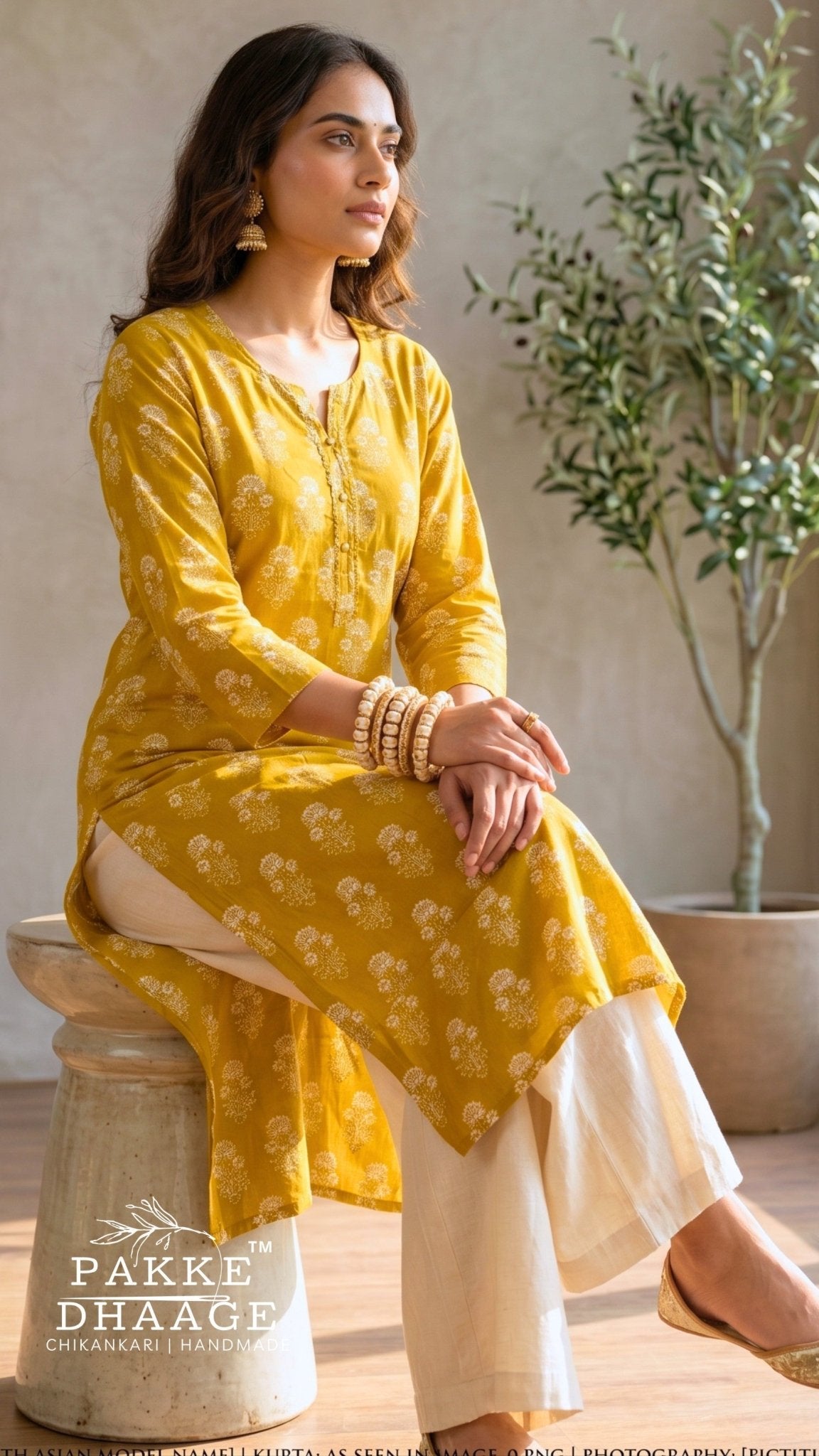 A woman models a yellow printed kurta with white pants, sitting on a stone pedestal in a rustic indoor setting with a potted plant in the background. The brand name 'PAKE DHAAGE' is visible as a watermark at the bottom left.