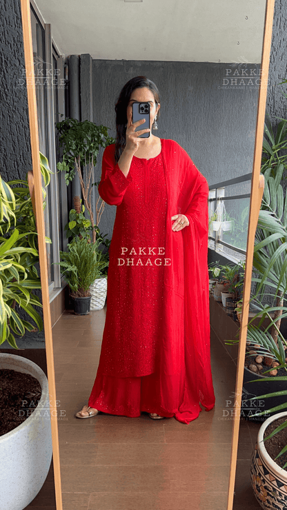 Person wearing a red chikankari suit set and a matching dupatta in an indoor setting with plants.