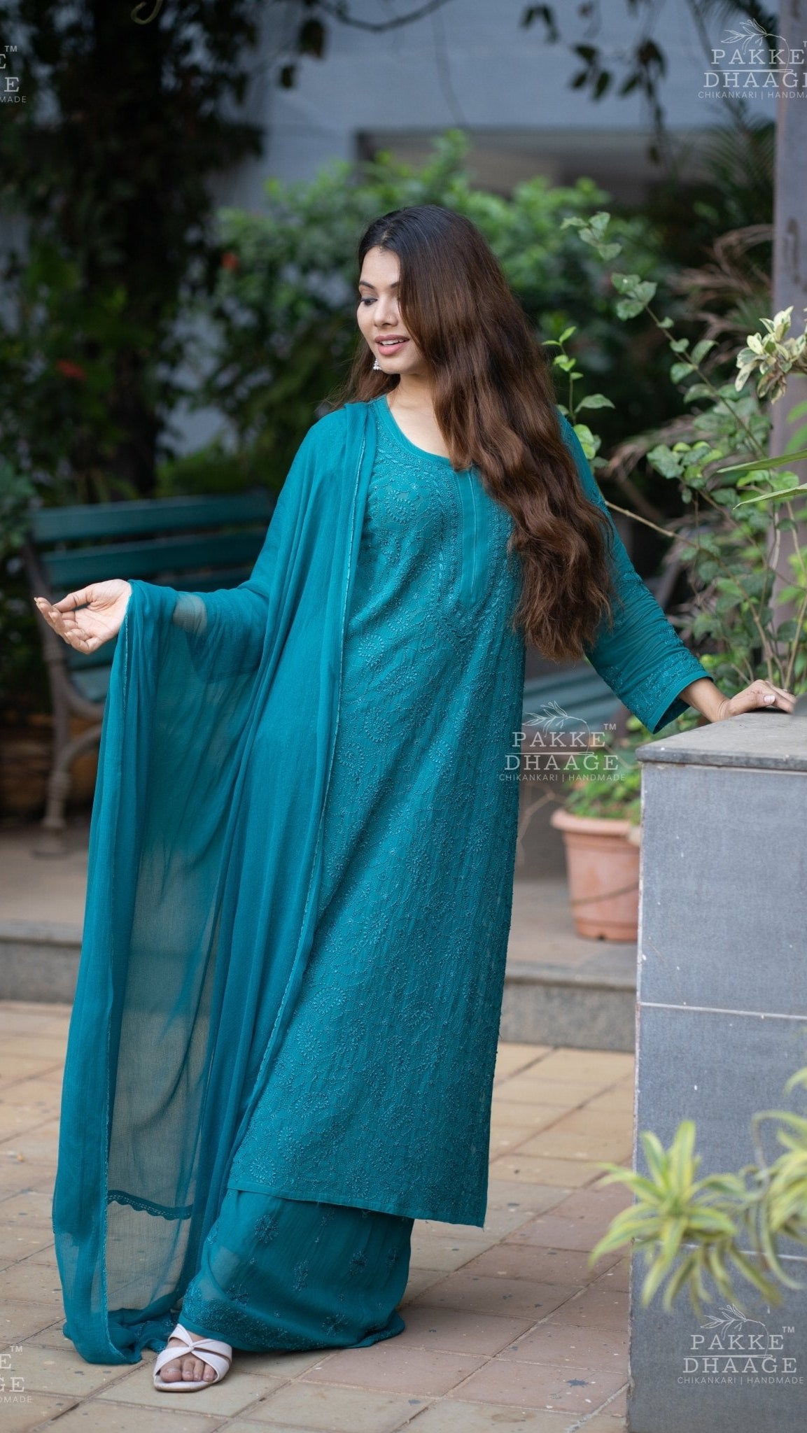 Woman in a teal Chikankari outfit standing outdoors with plants and furniture in the background